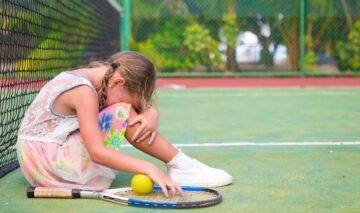 O fetiță tristă care stă cu capul pe genunchi pe un teren de tenis după ce a suferit o înfrângere