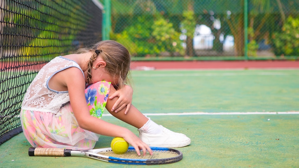 O fetiță tristă care stă cu capul pe genunchi pe un teren de tenis după ce a suferit o înfrângere