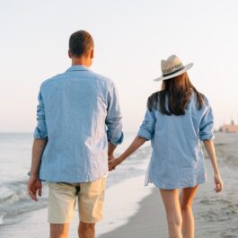 îndrăgostiți care se plimbă pe plajă și se țin de mână cu privirea către soare
