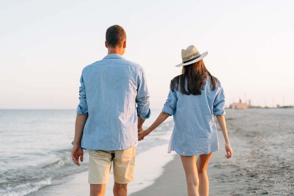 îndrăgostiți care se plimbă pe plajă și se țin de mână cu privirea către soare