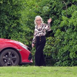 Angela Kelly în fața unei mașini roșii în timp ce le face cu mâna fotografilor