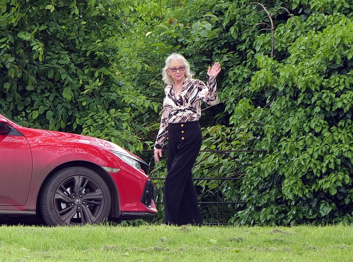 Angela Kelly în fața unei mașini roșii în timp ce le face cu mâna fotografilor