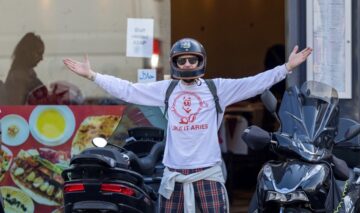 Paul Anderson cu o cască de motocicletă pe cap în timp ce pozează într-o ținută casual în fața unui magazin