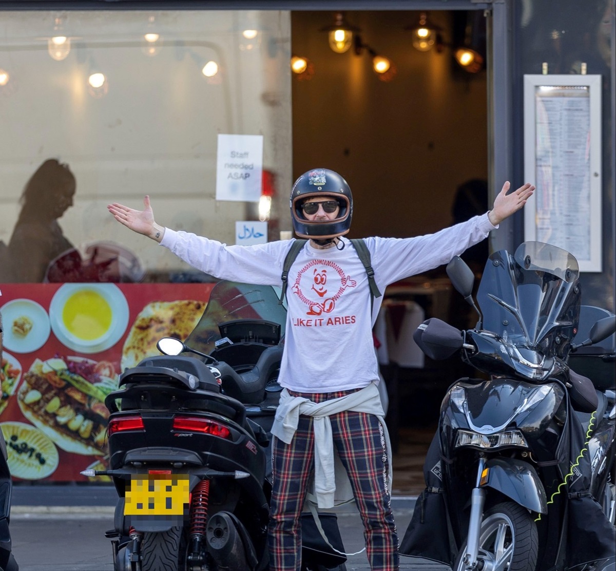 Paul Anderson cu o cască de motocicletă pe cap în timp ce pozează într-o ținută casual în fața unui magazin