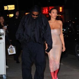 Kanye West într-o ținută all black în timp ce o ține pe Bianca Censori de mână, aceasta purtând o rochie transparentă din plastic și o pereche de cizme roșii