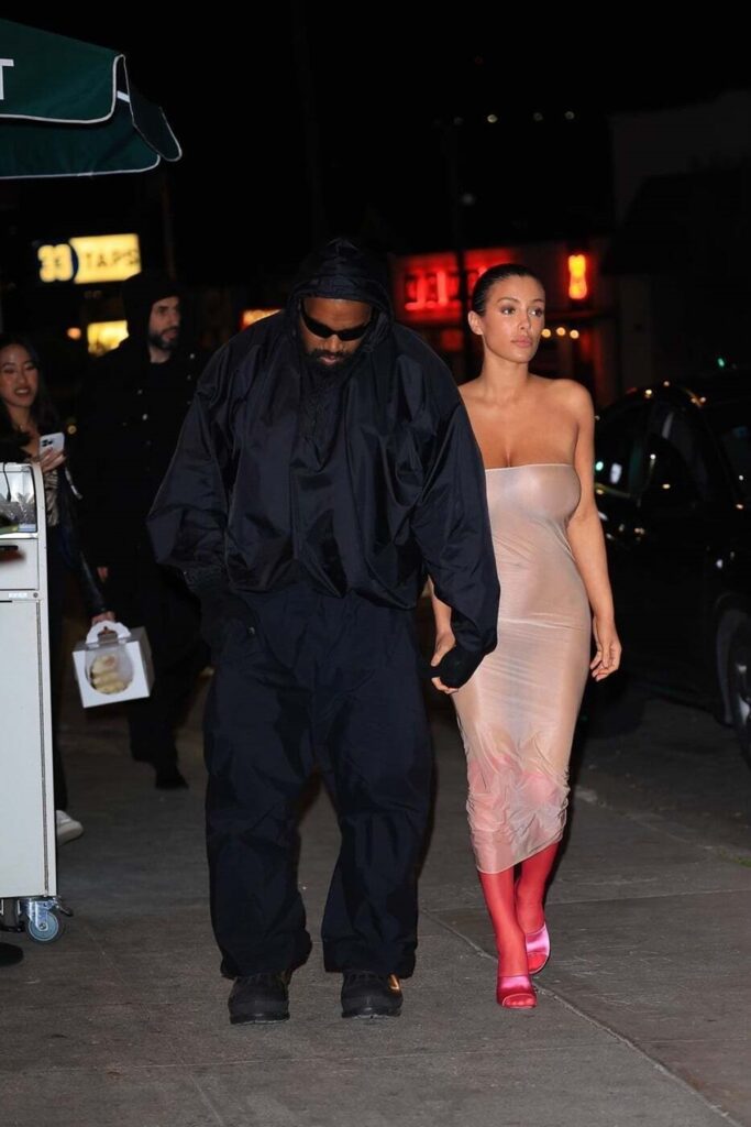 Kanye West într-o ținută all black în timp ce o ține pe Bianca Censori de mână, aceasta purtând o rochie transparentă din plastic și o pereche de cizme roșii