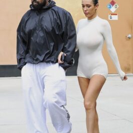 Kanye West într-o geacă neagră și o pereche de pantaloni albi alături de Bianca Censori care poartă un costum transparent