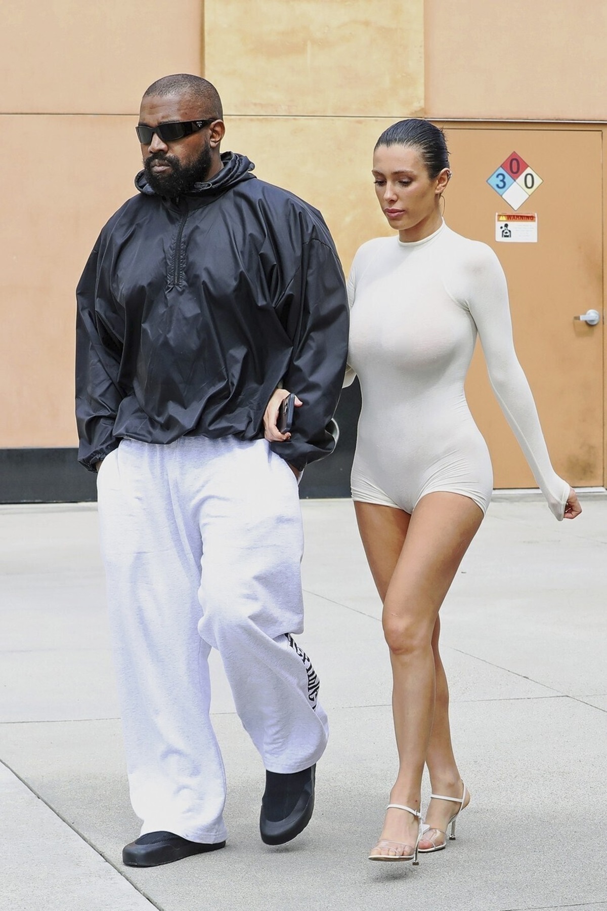 Kanye West într-o geacă neagră și o pereche de pantaloni albi alături de Bianca Censori care poartă un costum transparent