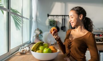 O femeie frumoasă care se află într-o bucătărie și ține în mână un măr în timp ce în fața sa are o fructieră cu banane, portocale și mere, fructe pe care e recomandat să le consumi dacă ai sindromul metabolic