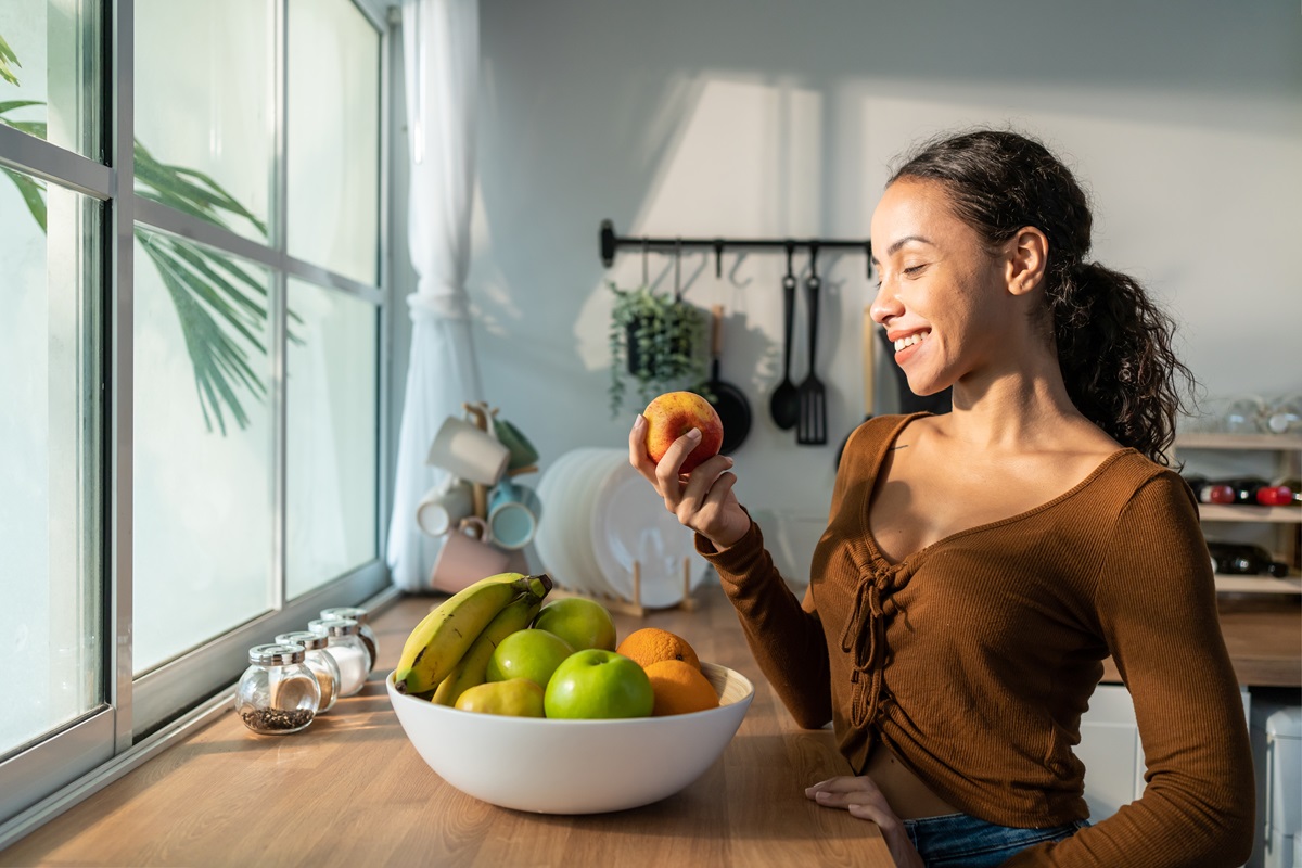 O femeie frumoasă care se află într-o bucătărie și ține în mână un măr în timp ce în fața sa are o fructieră cu banane, portocale și mere, fructe pe care e recomandat să le consumi dacă ai sindromul metabolic