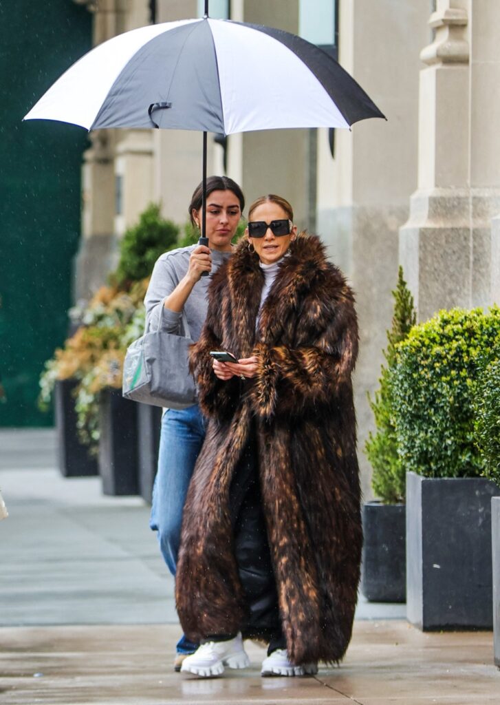 Jennifer Lopez în timp ce se plimbă pe stradă într-o haină de blană, iar asistenta ei ține o umbrelă