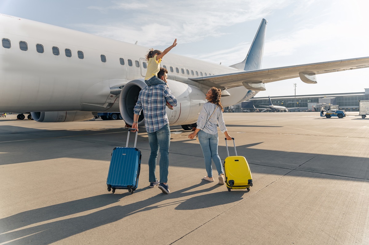 Un tată care ține un băiețel pe umeri alături de o mămică care duce o valiză galbenă și se îndreptă împreună spre avion