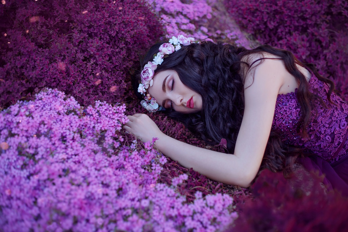 O femeie frumoasă care poartă o rochie mov și stă pe unpat de flori de liliac