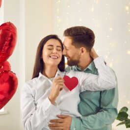 Portretul unui cuplu care se ține în brațe și se privesc în timp ce pe fundal se află mai multe baloane în formă de inimioară