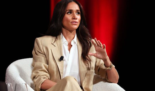 Meghan Markle poartă un costum deschis la culoare pe un fundal închis la culoare